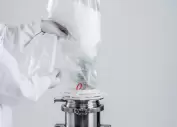 Operator in protective clothing connects a single-use transfer bag to a stainless steel container for the dust-free filling of white powder in a pharmaceutical or biotech application.