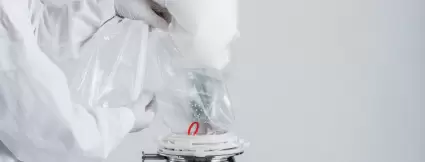Operator in protective clothing connects a single-use transfer bag to a stainless steel container for the dust-free filling of white powder in a pharmaceutical or biotech application.